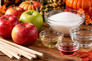 Fresh red and green apples beside bowls of sugar, corn syrup, water and red food colouring with wooden candy apple sticks.